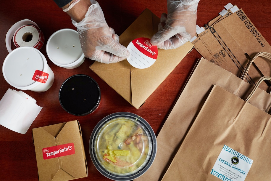 A person wearing gloves places a round “TamperSafe” sticker onto a brown paper food container, surrounded by takeout bags, food containers, and a roll of labels on a dark wooden table.