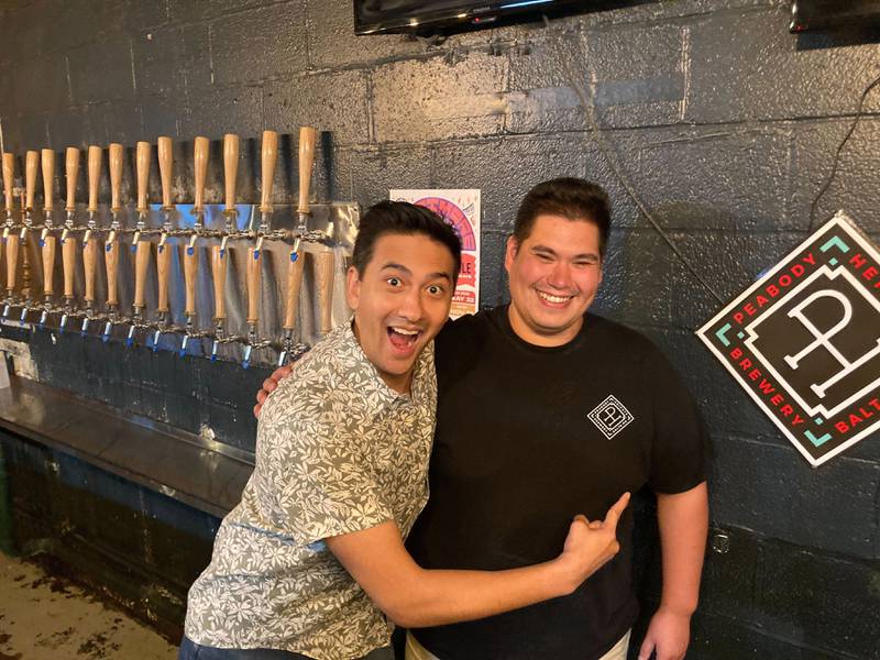 Two men smiling in front of a row of beer taps, one pointing at the Peabody Heights Brewery logo on the other’s t-shirt.