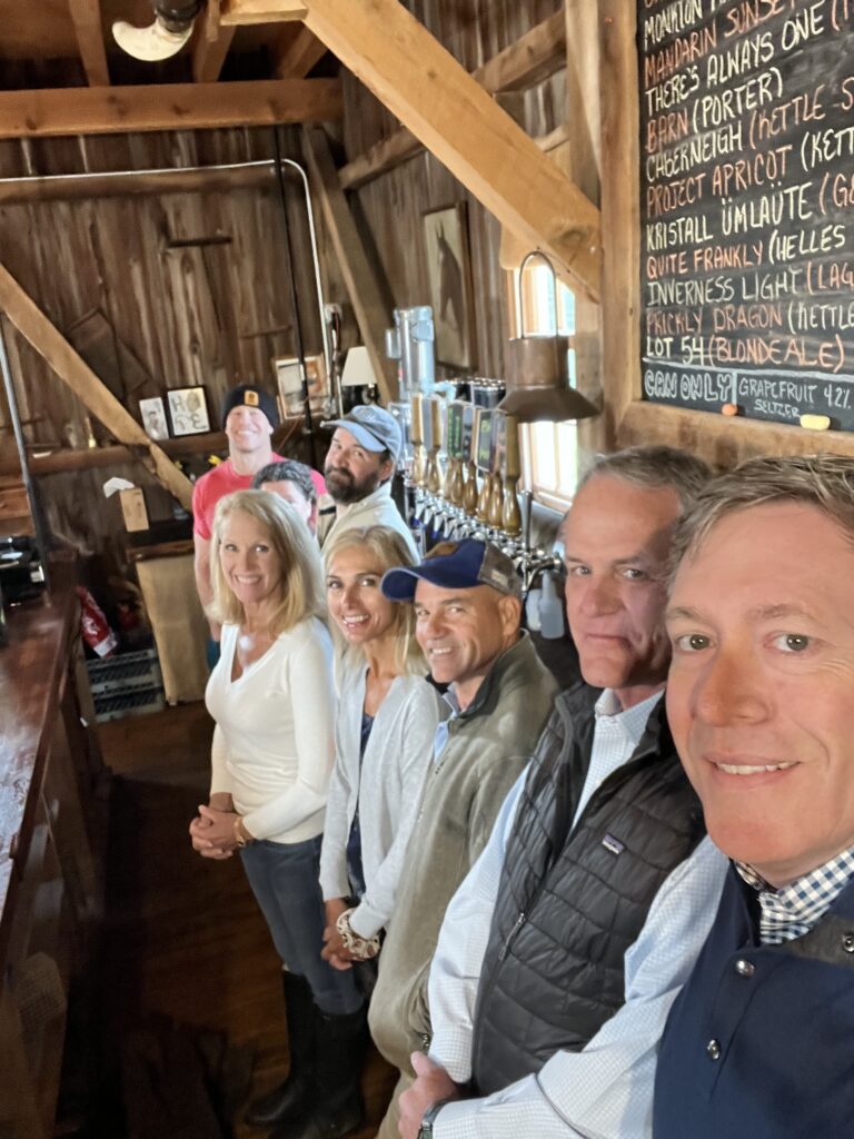 A group of seven people stand in a line smiling inside a rustic wooden barn brewery with a chalkboard menu of beers on the wall.