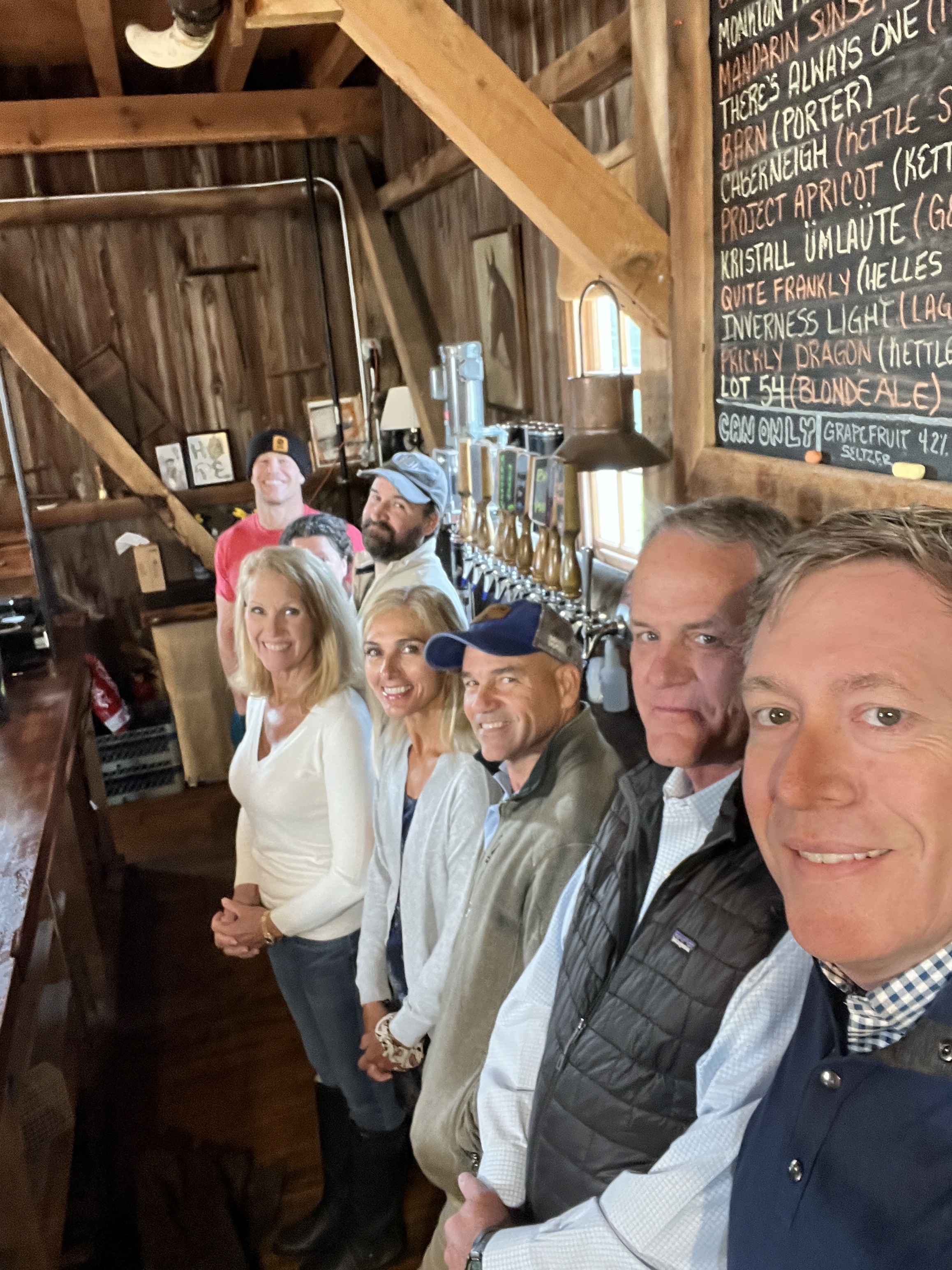 A group of seven people stand in a line smiling inside a rustic wooden barn brewery with a chalkboard menu of beers on the wall.