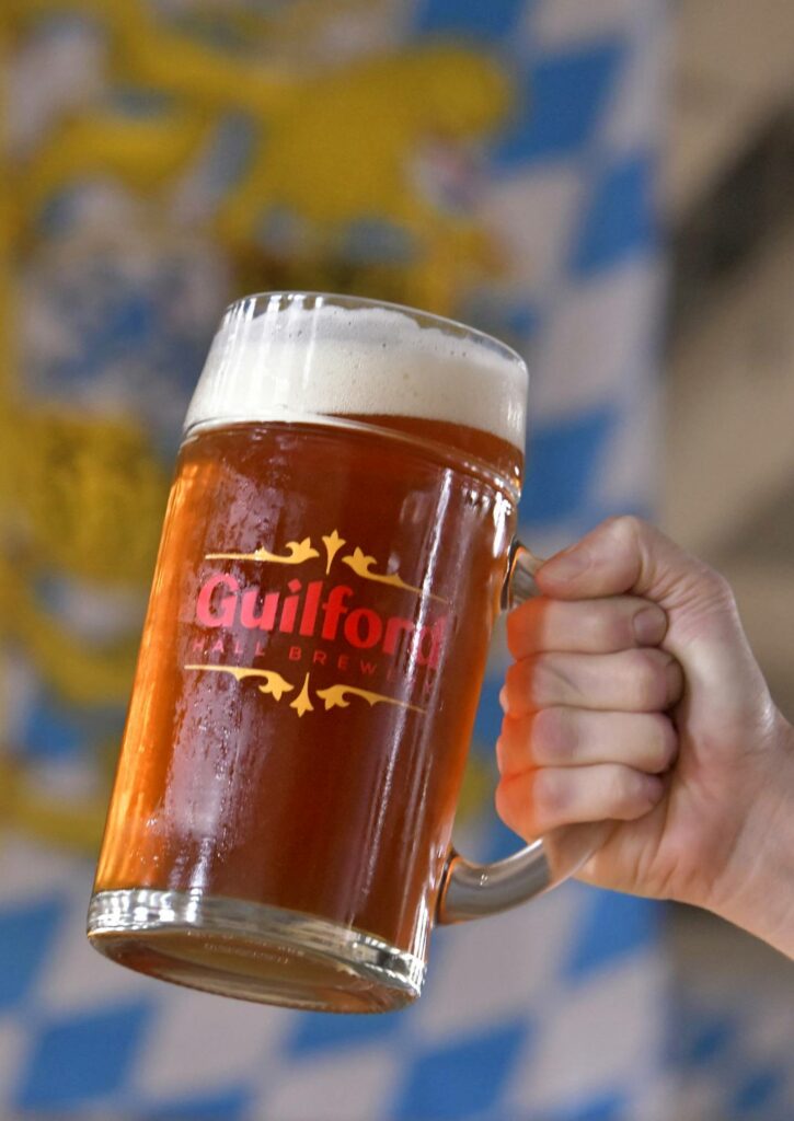 A hand holds a large glass beer mug with the Guilford Hall Brewery logo, set against a blue and white checkered background.