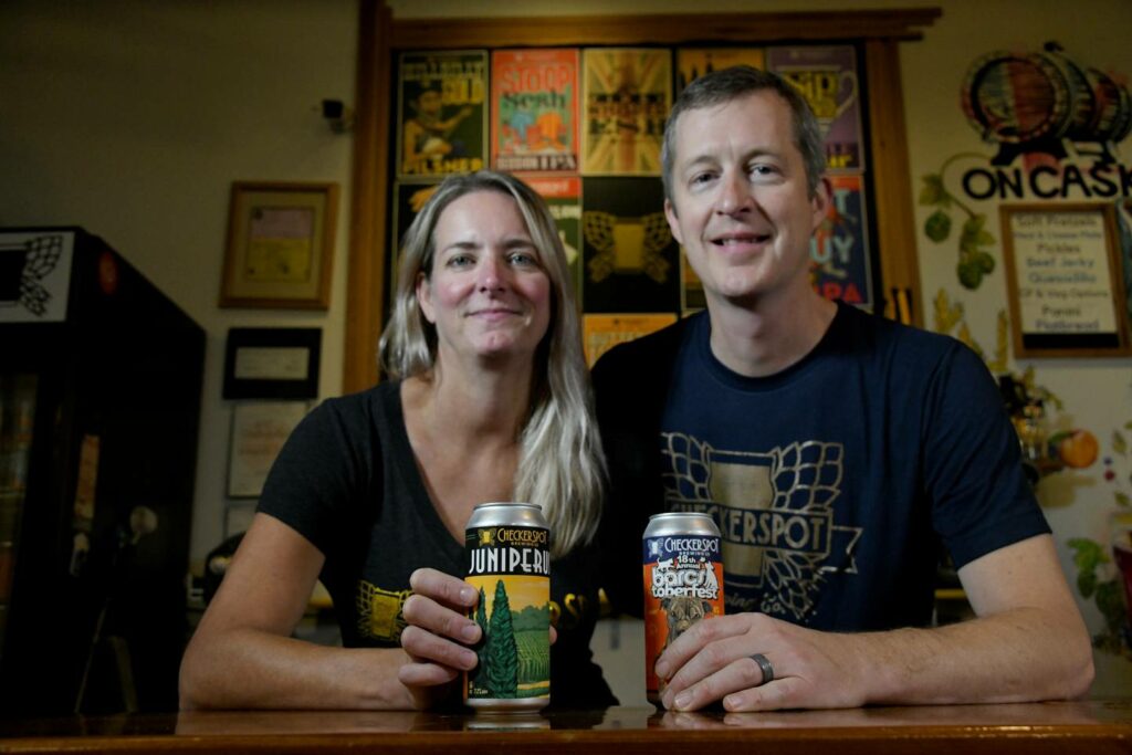 A man and woman standing behind a bar, each holding a can of Checkerspot beer.