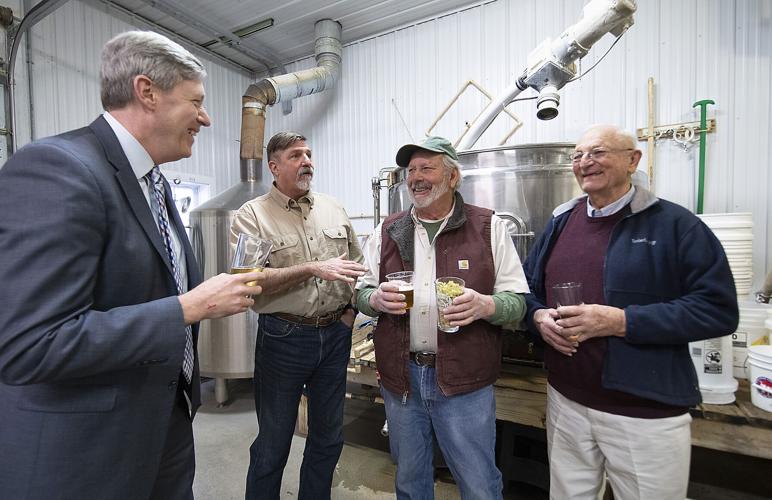 Three men stand in a brewery holding glasses of beer, with the man on the left in a suit speaking to the other two men.