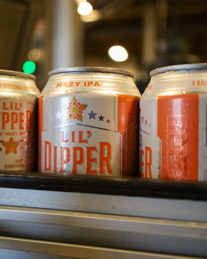 Three cold cans of Lil’ Dipper Hazy IPA beer lined up on a shelf.