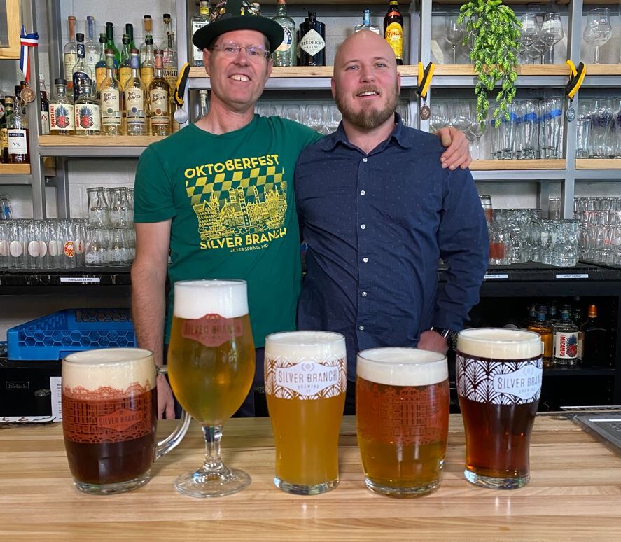 Two men stand behind a bar with five assorted glasses of beer on the counter in front of them.