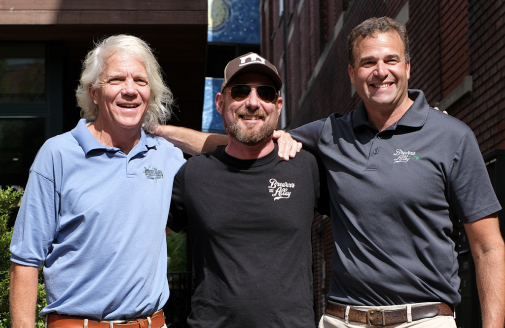 Three men wearing Brewers Alley branded apparel stand arm-in-arm and smile for the camera.