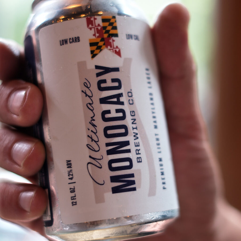A hand holding a can of Ultimate Monocacy Brewing Co. premium light Maryland lager.