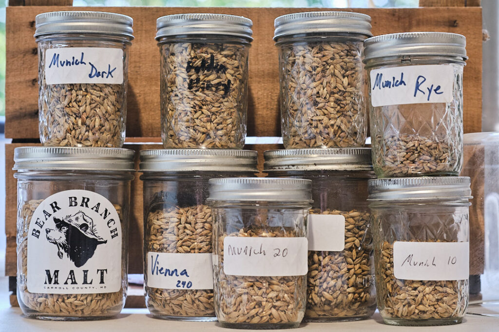 Eight mason jars filled with different types of brewing malt, each labeled with varieties including Munich, Vienna, and Rye, sitting in front of a wooden crate.
