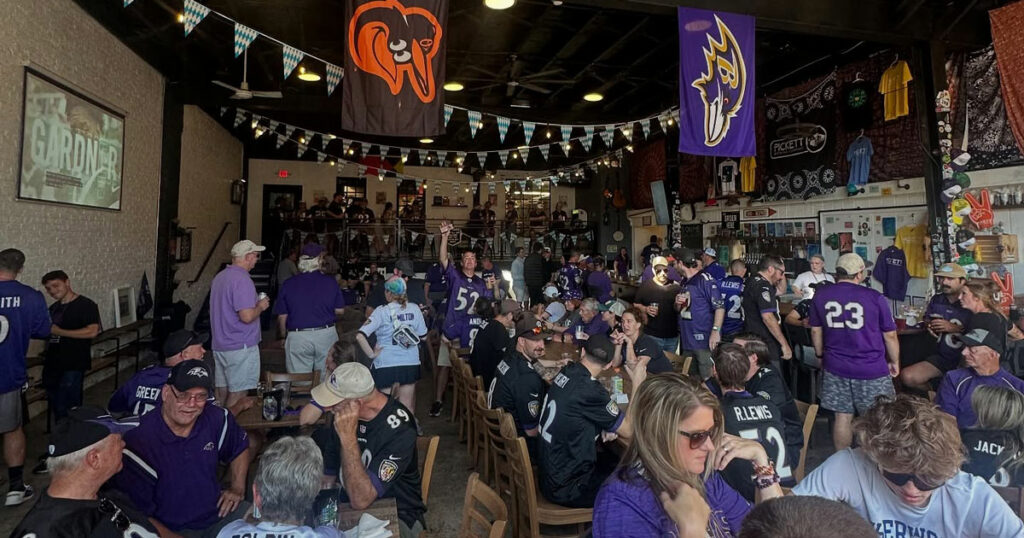 Patrons wearing Baltimore Ravens jerseys gather in a crowded sports bar decorated with team flags and banners.