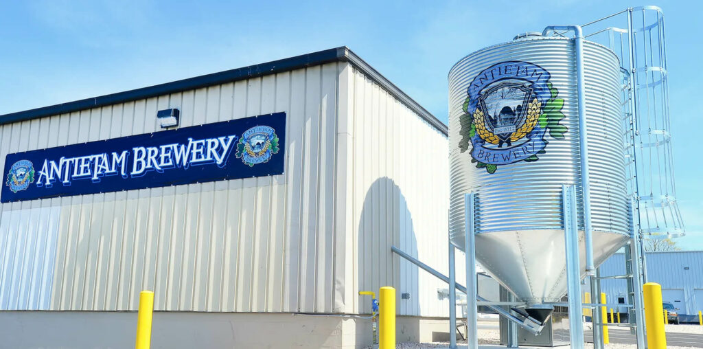 The exterior of Antietam Brewery with the company name and logo displayed on the metal building and a large silver grain silo.