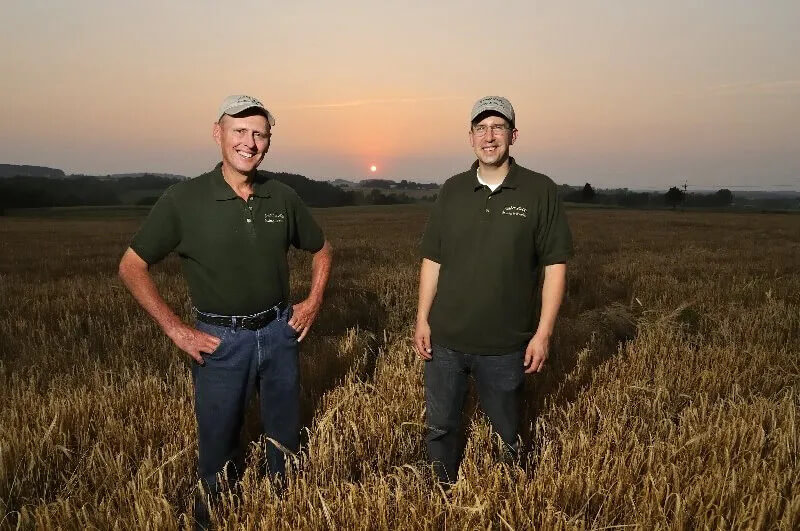 Two men in matching polo shirts and baseball caps standing in a wheat field at sunset.