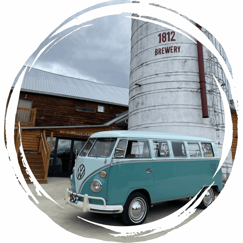 A teal and white vintage Volkswagen bus parked in front of a rustic wooden building and a silo labeled 1812 Brewery.