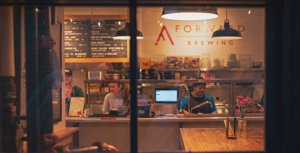 Interior view of Forward Brewing through a window, showing staff working behind a service counter with a menu board displayed on the wall above.