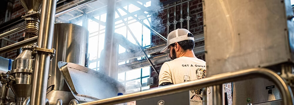 A brewer wearing a baseball cap works next to steaming stainless steel brewing equipment in a facility with brick walls.