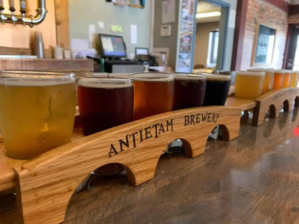 A wooden beer flight paddle labeled “Antietam Brewery” holding several glasses of beer of varying colors on a bar counter.
