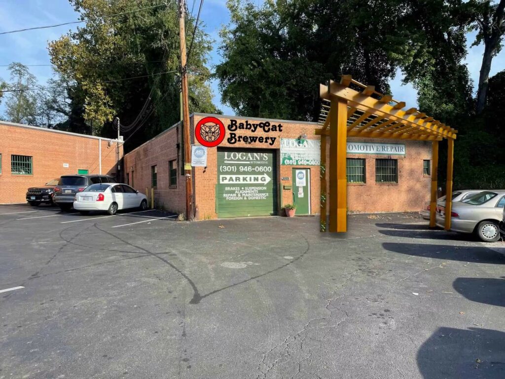 A brick building with signs for Baby Cat Brewery and Logan’s Automotive, featuring a large wooden pergola structure in the parking lot.