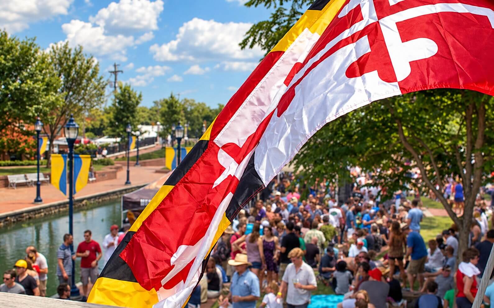 The Maryland state flag waving above a crowded outdoor festival along a canal.