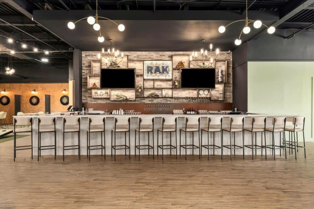 A long bar with stools facing a wall featuring the RAK logo, two television screens, and open shelving, with dartboards visible on the left.