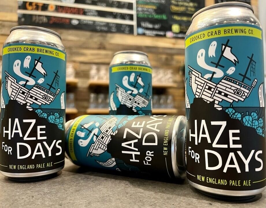 Three cans of Crooked Crab Brewing Co. Haze for Days New England Pale Ale, featuring a label illustration of a ghost ship, sitting on a counter in front of a menu board.
