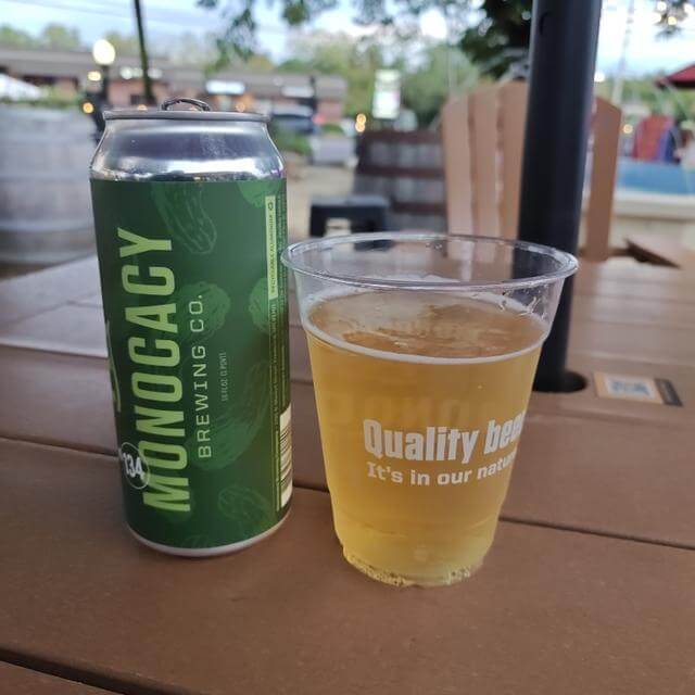 A can of Monocacy Brewing Co. beer next to a plastic cup filled with light-colored beer on a wooden table.