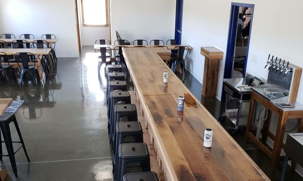 A long wooden bar with black metal stools and three cans of Upper Stem beer on the counter, with additional tables and chairs in a bright, modern taproom.