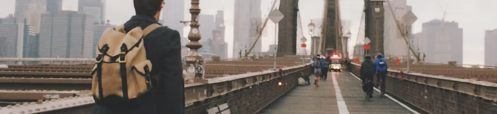 A person with a tan backpack walks along the Brooklyn Bridge with a city skyline in the background.