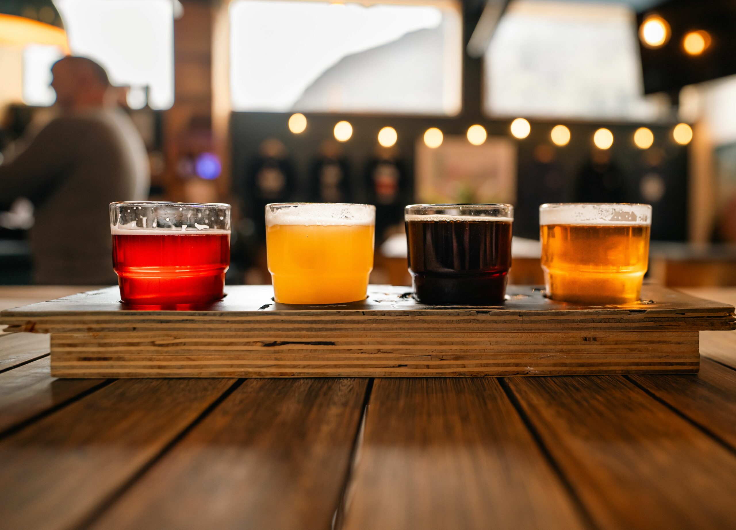 A flight of four different colored beers in small glasses on a wooden paddle sitting on a pub table.