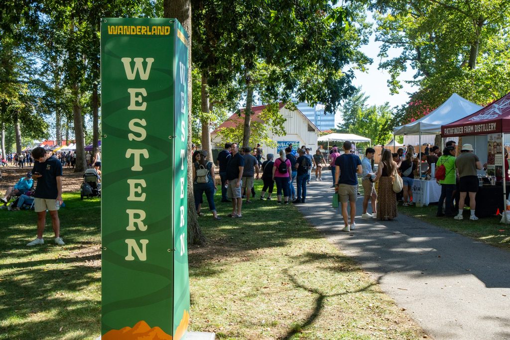 A tall green information sign labeled “WANDERLAND” and “WESTERN” stands at the entrance of a park festival featuring vendor booths and people walking along a path.