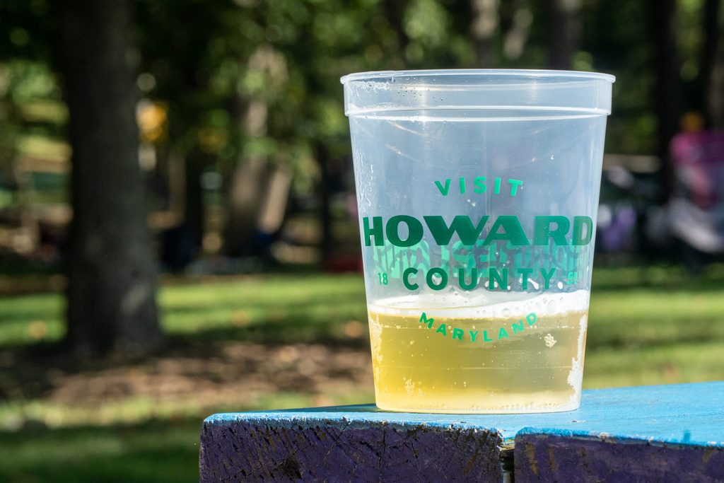 A plastic cup partially filled with a light-colored beverage, featuring the text “Visit Howard County Maryland” in green, sitting on a blue wooden surface outdoors.